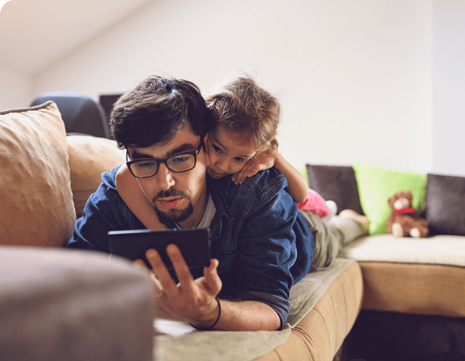 Man laying on his stomach on the couch looking at a tablet with his daughter on his back looking over his shoulder.