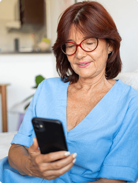 A senior woman with red hair and glasses, wearing a blue shirt, looks at her smartphone while sitting comfortably at home.