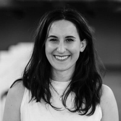 A black-and-white photo of Manya Ellenberg with long dark hair, smiling warmly while wearing a light-colored sleeveless top