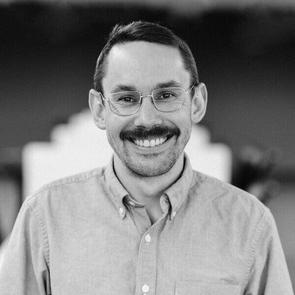 Black-and-white headshot of Scott Voigt smiling and wearing glasses and a button-down shirt, with a blurred background