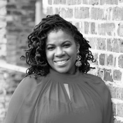 Stephanie B. Broussard leaning against a brick wall and smiling at the camera.